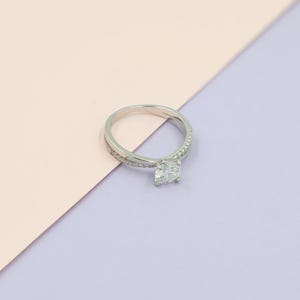 Pode incluir: Um anel de prata com uma grande pedra preciosa clara de corte quadrado como ponto focal. A banda divide-se em duas mechas, uma das quais é embelezada com pequenas pedras claras. O anel está sobre um fundo rosa pastel e roxo.