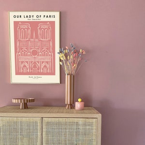 May include: A framed print of Notre Dame de Paris with the text "Our Lady of Paris" and "Ecole de France 1163-1345". A vase with pink and brown stripes holds dried flowers in shades of pink, yellow, and purple. A pink ceramic container with a yellow knob sits on a wooden cabinet with a woven rattan front.