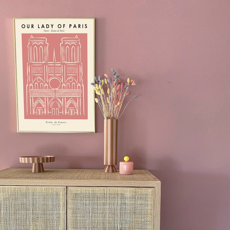 May include: A framed print of Notre Dame de Paris with the text "Our Lady of Paris" and "Ecole de France 1163-1345". A vase with pink and brown stripes holds dried flowers in shades of pink, yellow, and purple. A pink ceramic container with a yellow knob sits on a wooden cabinet with a woven rattan front.