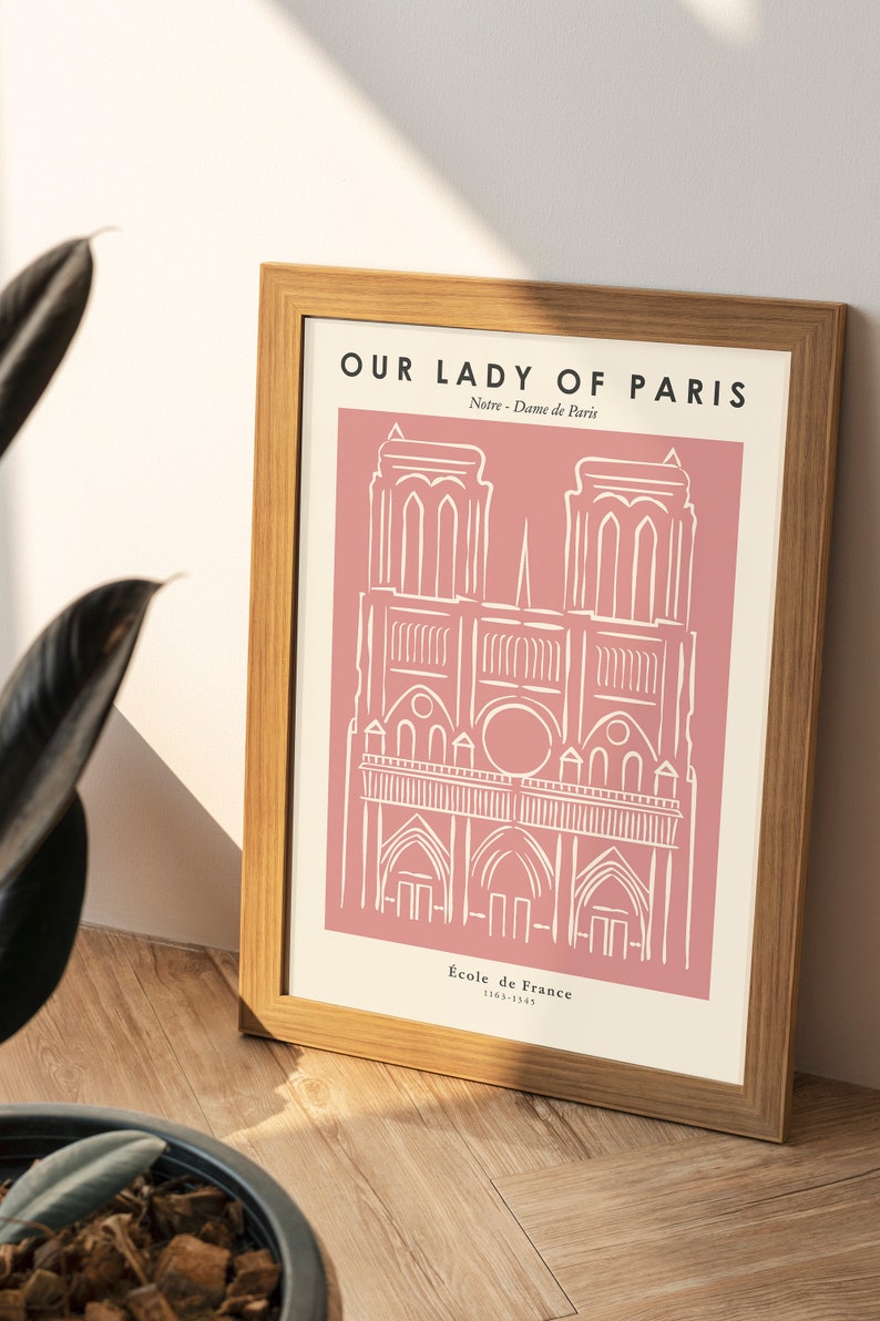 May include: A framed print with a pink background and a white line drawing of Notre Dame Cathedral in Paris. The text "Our Lady of Paris" is at the top of the print, and the text "Notre Dame de Paris" is below the drawing. The text "Ecole de France 1163-1345" is at the bottom of the print.