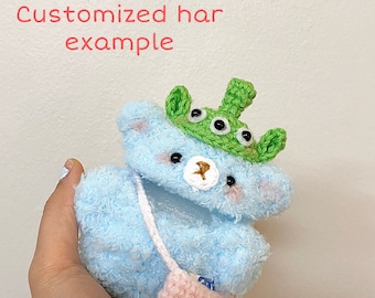 May include: A light blue teddy bear plushie wearing a green alien hat with three eyes and a pink crocheted purse. The text "Customized har example" is visible in the image.