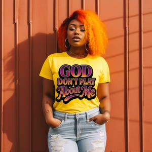 May include: Yellow t-shirt with the text "GOD DON'T PLAY ABOUT ME" in a purple and black design. The person is wearing light blue jeans and has orange hair.