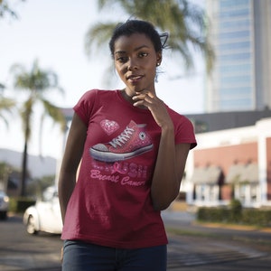 May include: A burgundy t-shirt featuring a graphic of a pink high-top sneaker with a diamond and the text "Crush Breast Cancer". The design includes a pink ribbon, a symbol of breast cancer awareness.
