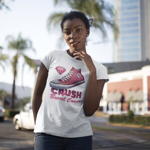 May include: White t-shirt with a pink high-top sneaker graphic, a pink heart-shaped diamond, and the text "Crush Breast Cancer" in pink. The shirt is a crew neck and has short sleeves.