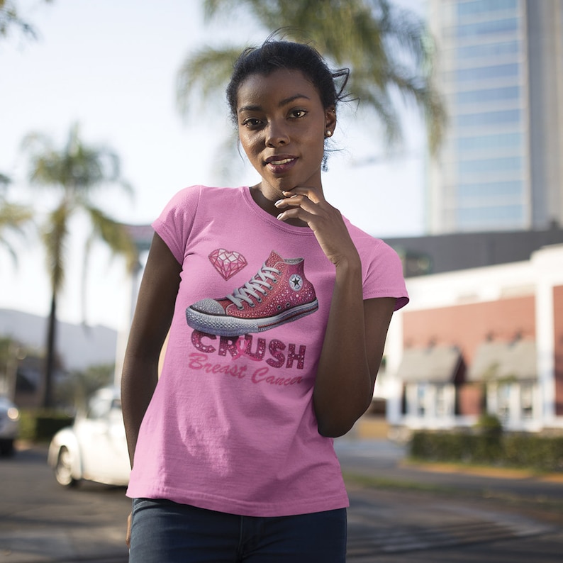 May include: Pink t-shirt featuring a sparkly red high-top sneaker graphic with a diamond and the text "Crush Breast Cancer." The shirt is a solid pink color and has short sleeves.