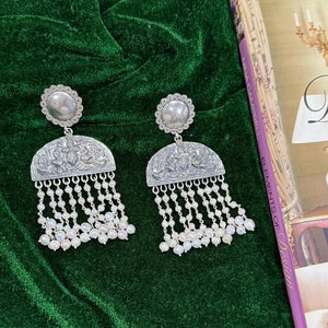 May include: Silver-coloured earrings with detailed engraved patterns. Each earring has a circular top, a detailed half-moon pendant, and multiple strands of small, white pearls. The earrings are displayed on a dark green velvet surface.