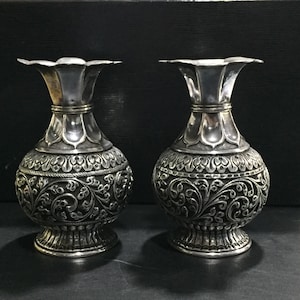Op de afbeelding: Twee zilverkleurige vazen met ingewikkelde bloempatronen. De vazen hebben een bolvormige body, een uitlopende bovenkant en een gedetailleerde basis. Het ontwerp omvat reliëf bloemen en krullen, waardoor een vintage esthetiek ontstaat. De vazen worden tegen een donkere achtergrond getoond.