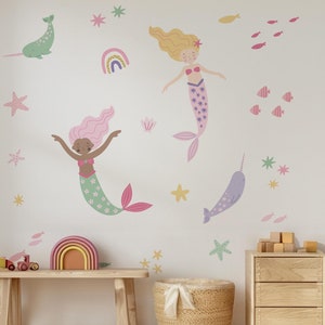 Può includere: Un muro bianco con una varietà di adesivi murali colorati, tra cui sirene, narvali, stelle e un arcobaleno. Gli adesivi sono disposti in uno schema fantasioso e giocoso.