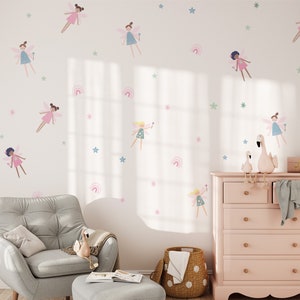 Könnte beinhalten: Eine weiße Wand mit rosa und blauen Feenstickern, eine rosa Kommode mit sechs Schubladen, ein grauer Sessel mit einer Wolldecke, ein grauer Hocker und ein Weidenkorb.