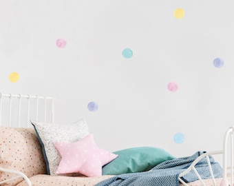 Puede incluir: Una cama blanca con una funda nórdica de lunares rosa y blancos y una manta de color azul y blanco. Hay varias pegatinas de lunares de colores en la pared detrás de la cama.