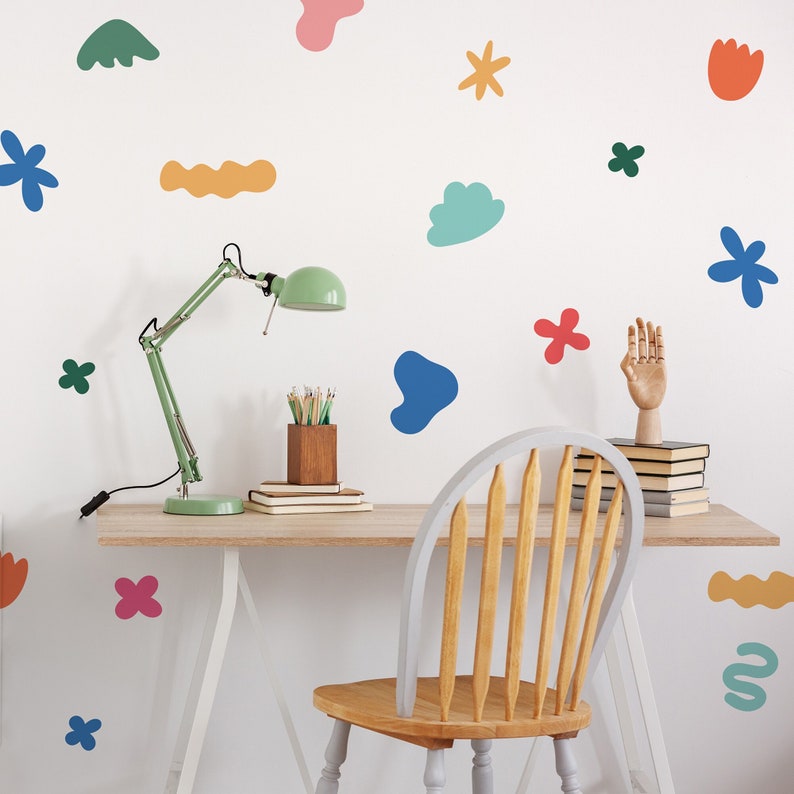 May include: A white wall decorated with colourful abstract shapes and a green desk lamp. A wooden chair sits in front of a desk with books and pencils.