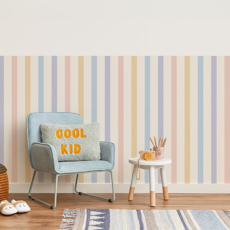 May include: A child's room with a light blue chair, a small white table, and a striped rug. The chair has a pillow with the words "COOL KID" in orange. The wall has vertical stripes in pastel colors.