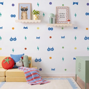 May include: A wall decal set featuring repeating patterns of blue superhero masks, teal lightning bolts, and multicoloured dots in orange, green, and purple. The decals are applied to a white wall above a yellow sofa with a tomato-shaped pillow.