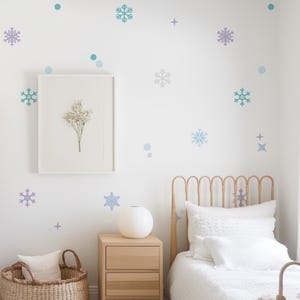 May include: A white wall decorated with blue and purple snowflake and circle decals. A white bed with a wooden headboard and white bedding is in the bottom right corner of the image. A white picture frame with a dried flower arrangement is on the wall. A small wooden bedside table with drawers is to the left of the bed.