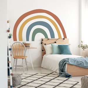 May include: A white bedroom with a wooden bed, a rainbow wall decal, and a white desk with a wooden chair. The bed has a white duvet, a blue blanket, and a few pillows. There are string lights hanging above the bed.