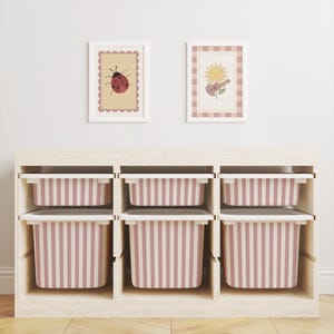 May include: A light wood storage unit with six removable bins featuring vertical dusty pink and white stripes. Above the unit, two framed wall prints are displayed: one with a ladybug illustration and one with a sunflower playing a guitar.