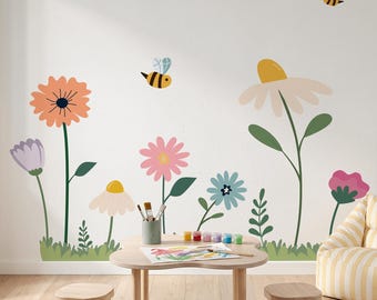 May include: A wall decal set featuring colorful flowers in shades of orange, pink, blue, and white, accompanied by green stems, leaves, and grass. Two small yellow and black bees are depicted flying above the floral arrangement on the wall.
