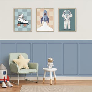 Op de afbeelding: Drie ingelijste wandkunstprints met een ruimtethema. Eén toont een rolschaats, een andere een lancerende raket en de derde een astronaut. De prints bevinden zich in een kinderkamer met een blauwe muur en een kleine stoel.