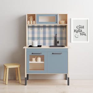 May include: A blue and white play kitchen with a black stovetop, sink, and cabinet doors. The kitchen has a checkered backsplash and a sign that reads "Be Cool Be Kind Be You".