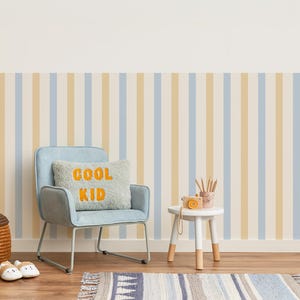 May include: A child's room with a light blue chair, a small white table, and a pillow that says "COOL KID" in orange letters. The wall has vertical stripes in light blue, beige, and white. A small rug and a basket are on the wooden floor.