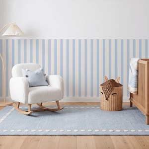 Peut inclure: Un fauteuil à bascule blanc texturé avec un coussin bleu clair, un tapis bleu clair à bordure pompons blancs, un panier de rangement tressé en forme de renard et un lit à barreaux en bois avec une couverture, devant un mur à rayures verticales.