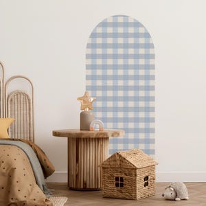 May include: A light blue and white gingham pattern wall decal in an arched shape. A wooden side table with a star-shaped lamp, a small rainbow, and a woven house are in front of the wall decal. A bed with a brown comforter is on the left.
