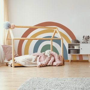May include: A wooden toddler bed with a rainbow wall decal in shades of red, yellow, blue, and green. The bed has a white blanket and a pink knitted throw with a stuffed animal on top.