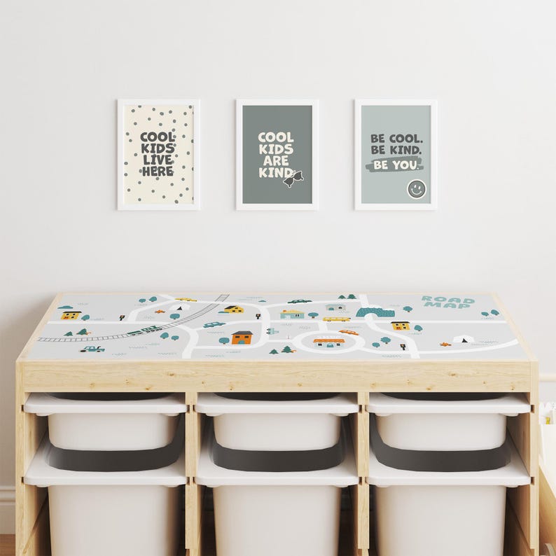 May include: A wooden play table with a gray and white road map design on the top. The table has three white storage bins underneath. Three framed prints with inspirational messages hang on the wall behind the table. The prints say "Cool Kids Live Here", "Cool Kids Are Kind", and "Be Cool. Be Kind. Be You."