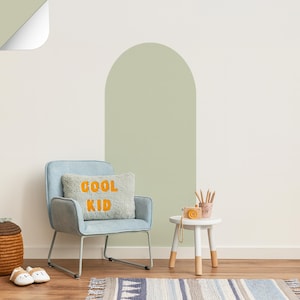 Può includere: Una poltrona azzurra con un cuscino con la scritta "COOL KID" in arancione. Un tavolino bianco con una macchina fotografica giocattolo e matite. La stanza ha un tappeto, un cesto intrecciato e un muro con un arco dipinto.