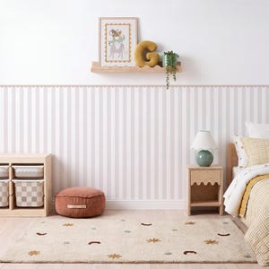 May include: A bedroom featuring light pink and white striped wallpaper, a wooden shelf with a framed animal print, a yellow letter G pillow, and a green plant. Includes a wooden nightstand, a lamp, a terracotta floor cushion, and a patterned rug.