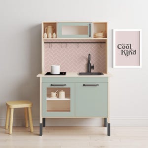 May include: A light blue and wood play kitchen with a black sink and stovetop. The kitchen has a pink and white tiled backsplash and a shelf with a white pot and a white mug. The kitchen has a sign that says "It's Cool to be Kind".