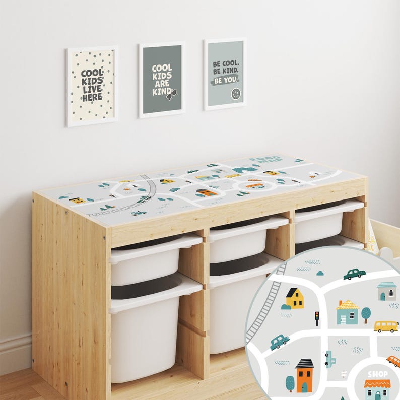 May include: A wooden toy storage unit with three white bins and a removable playmat featuring a city street scene with cars, houses, and a shop. The playmat is gray with blue and green accents. The unit has a light wood finish.
