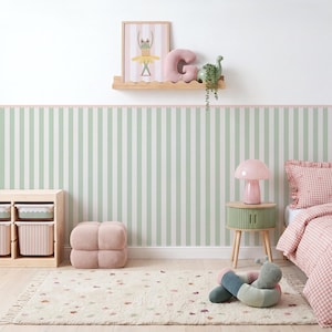 Può includere: Una camera da letto con carta da parati a righe verde salvia e bianco, una mensola in legno chiaro con stampa di rana, un cuscino a forma di lettera G rosa e un dinosauro verde. Include un mobile contenitore, una lampada a fungo e un letto a quadri.