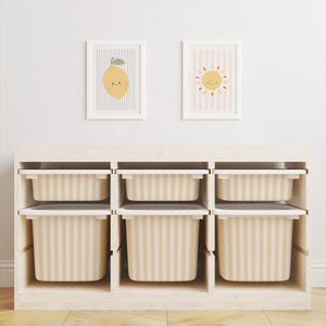 Könnte beinhalten: Ein helles Holzregal mit sechs Fächern, in denen sich jeweils eine gestreifte beige-weiße Box befindet. Zwei gerahmte Drucke einer Zitrone und einer Sonne hängen darüber. Der Raum wirkt hell und luftig.