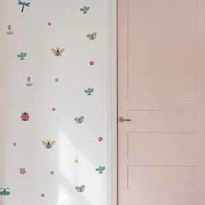 Op de afbeelding: Een witte muur versierd met kleurrijke insecten- en bloemenstickers. De stickers bevatten vlinders, bijen, lieveheersbeestjes, libellen en bloemen in roze, blauw en geel. Rechts is een lichtroze deur.