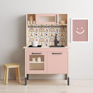 May include: A pink toy kitchen playset with a black countertop, two cabinets, and a sink. The backsplash features a colorful floral and rainbow pattern. A small wooden stool is in the foreground.