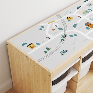 May include: A wooden toy storage unit with three drawers and a removable top. The top features a gray and white printed design of a town with houses, cars, trees, a train, and a tractor.