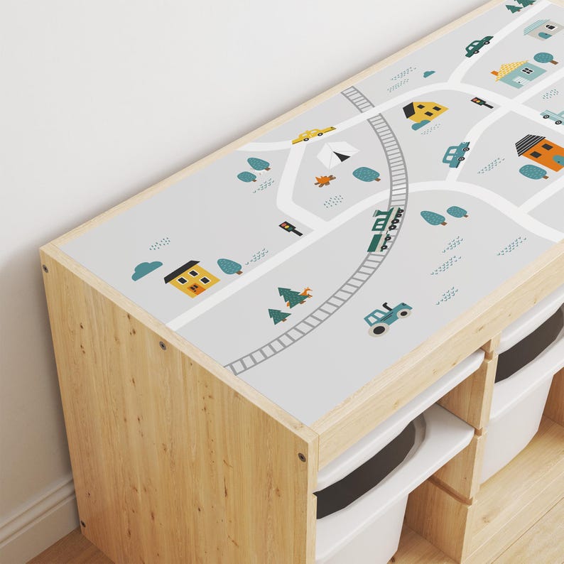 May include: A wooden toy storage unit with three drawers and a removable top. The top features a gray and white printed design of a town with houses, cars, trees, a train, and a tractor.