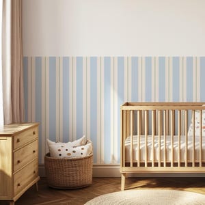May include: A nursery interior with a light wood cot, a chest of drawers, and a wicker basket holding pillows. The wall is decorated with vertical stripes in pale blue and beige. A round rug is on the wooden floor.
