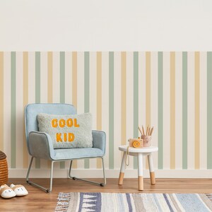 May include: A child's room with a light blue chair, a small white table, and a striped wallpaper in pastel colors. A pillow with "COOL KID" in orange letters sits on the chair. A small camera and pencils are on the table. A rug is on the floor.