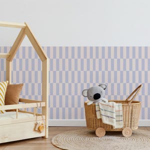 Op de afbeelding: Een licht houten bed met een geel en bruin kussen. Een rieten mand op wielen bevat een koala knuffel en een gestreepte deken. De achtergrond heeft blauw en beige gestreept behang en een rond geweven vloerkleed.