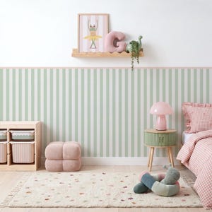 May include: A bedroom featuring sage green and white striped wallpaper, a light wood shelf with a frog print, a pink letter G pillow, and a green dinosaur. Includes a light wood storage unit, a pink mushroom lamp, and a pink gingham bed.