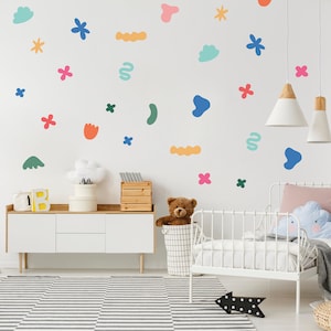 May include: A white wall decorated with colourful abstract shapes and flower stickers. A white dresser with a teddy bear in a wire basket sits in front of the wall. A white cot with a pink pillow and a blue cloud-shaped pillow is in the foreground.