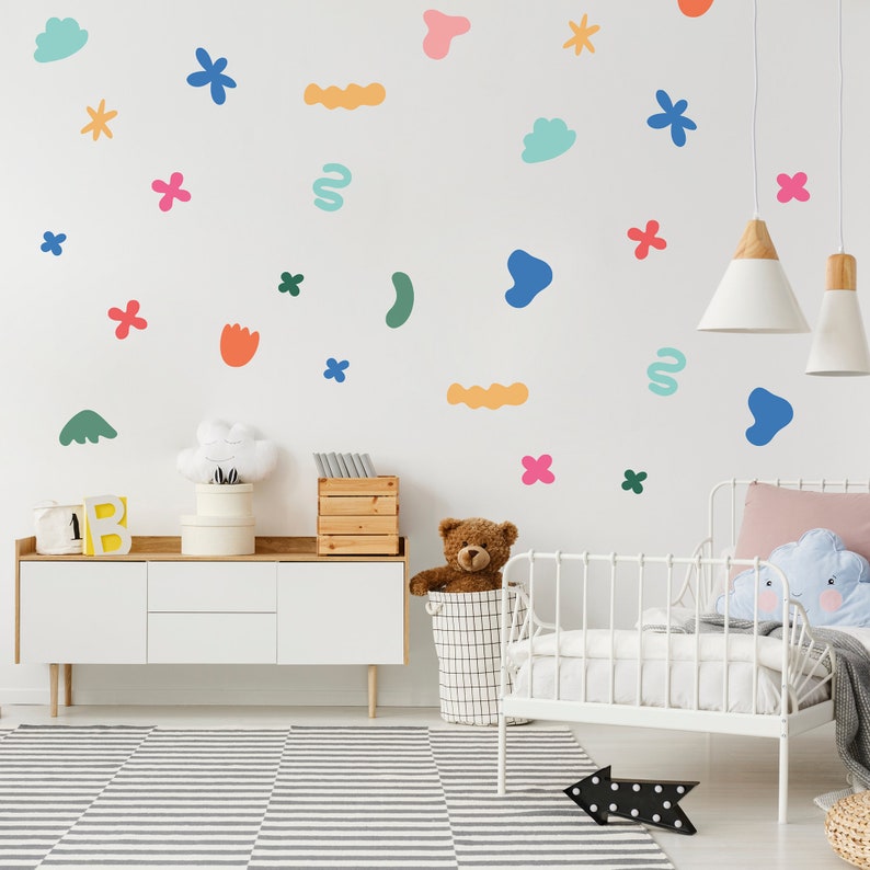 May include: A white wall decorated with colourful abstract shapes and flower stickers. A white dresser with a teddy bear in a wire basket sits in front of the wall. A white cot with a pink pillow and a blue cloud-shaped pillow is in the foreground.