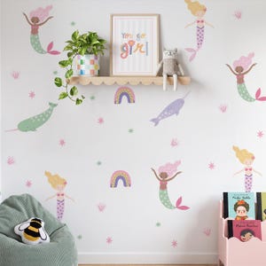May include: A wall decal set featuring mermaids, narwhals, rainbows, and stars in pastel colours. A framed print with the text You go girl sits on a scalloped wooden shelf above a green beanbag chair and a pink book storage bin with books.