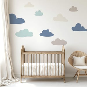May include: A nursery scene featuring a wooden crib with a white mattress and a wicker chair with a white pillow. The white wall is decorated with various pastel-colored cloud decals in shades of blue, gray, and beige.
