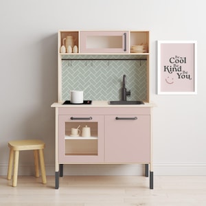 May include: A pink and wood children's play kitchen with a black stovetop, sink, and cabinet hardware. The kitchen has a small shelf above the stovetop and a larger shelf above the sink. The kitchen has a sign that says "Be Cool Be Kind Be You".
