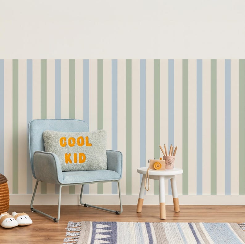 May include: A child's room with striped wallpaper in blue, green, and cream. A light blue chair holds a pillow with "COOL KID" in orange. A small white table with wooden legs holds a toy camera and pencils. A striped rug and wooden floor complete the scene.