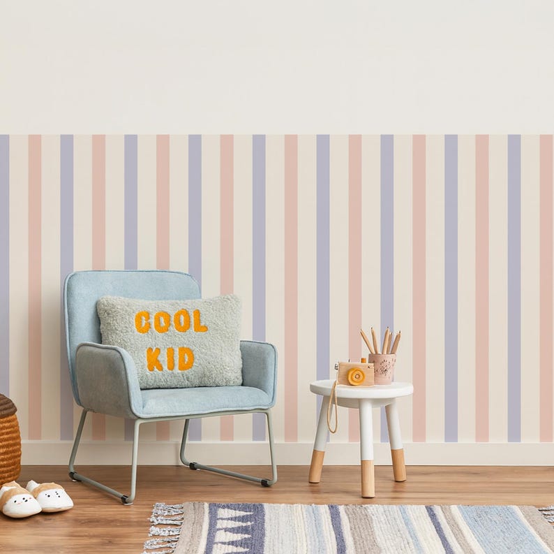 May include: A child's room with a light blue chair, a small white table, and a striped wallpaper in pastel colors. A pillow with "COOL KID" in orange letters sits on the chair. A rug and a basket are on the wooden floor.