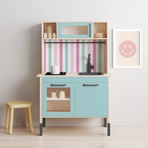 May include: A light blue toy kitchen playset with a black sink, stovetop, and oven. The kitchen has a pink and blue striped backsplash and a shelf with a white pot and two white mugs. A wooden stool is in the foreground.
